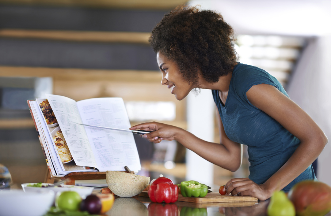 A woman enthusiastically looks at a cookbook with healthy ingredients all around her.