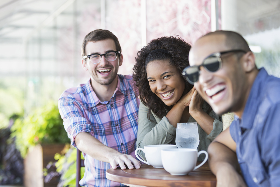 Younger group of friends smiling and having coffee at an outside table.